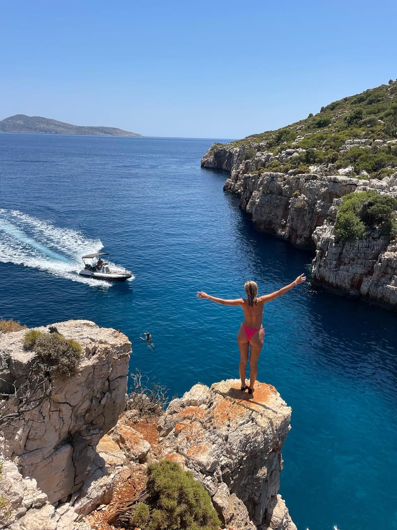 A guest with arms raised on the cliff above the inlet, the boat visible below